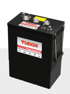 Black car battery with a Tudor label, two top terminals and a carrying handle, resting on a light gray surface.